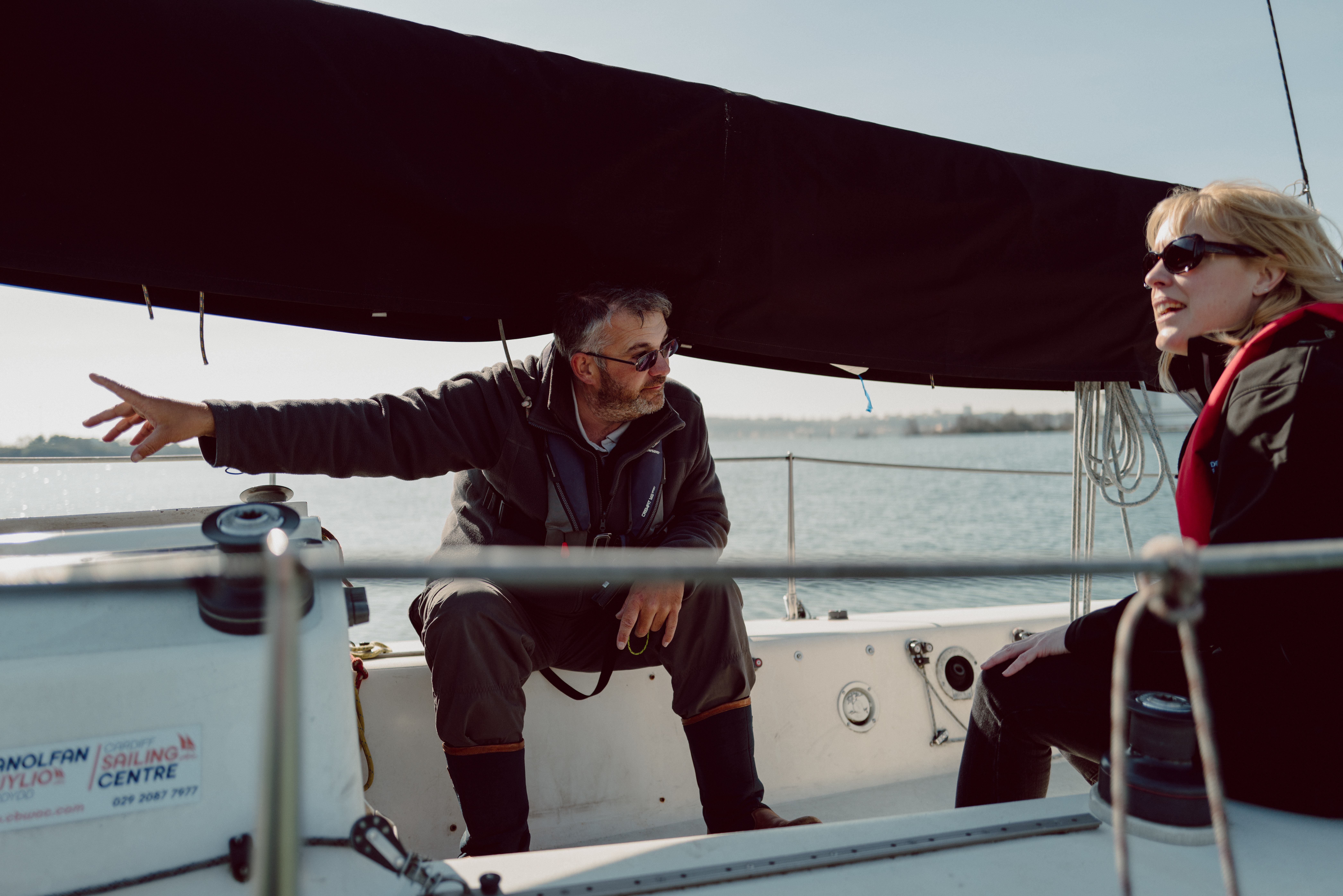 Two people sat on a sailing boat. One is pointing,